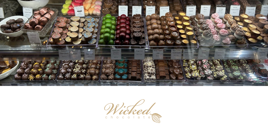 Wicked Chocolate logo sits underneath rows of fresh chocolates behind a display counter.